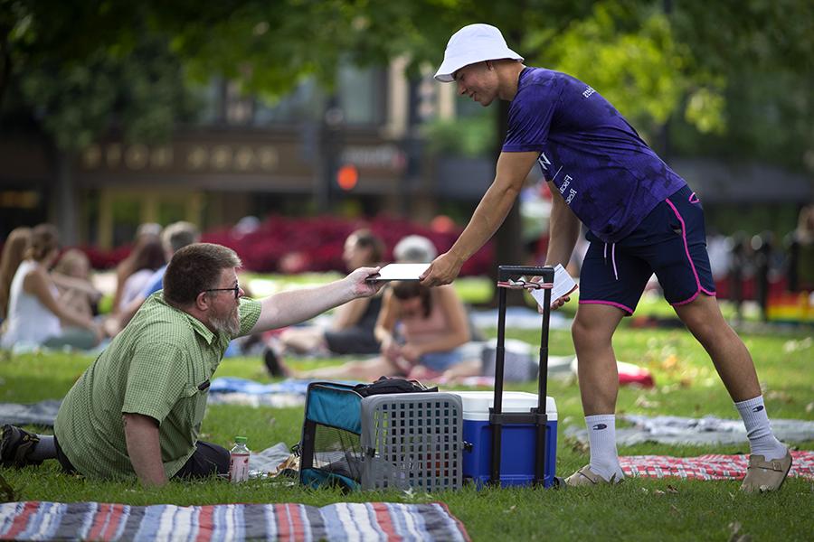 一位身穿紫色衬衫、头戴白色帽子的Economics专业学生将一份调查报告递给坐在公园草坪椅上的人.
