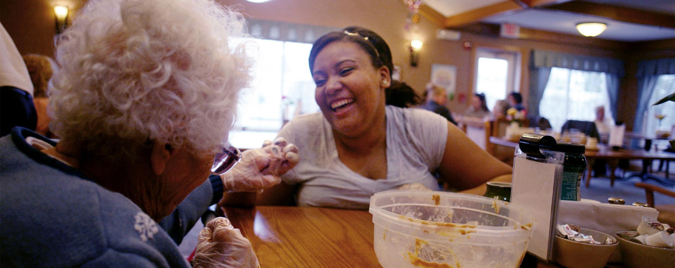 A student smiles and laughs with an elderly woman and holds her hand.