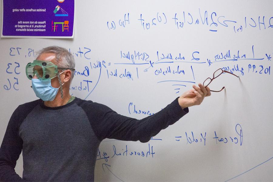 A faculty member points at the whiteboard while wearing safety goggles and a mask.