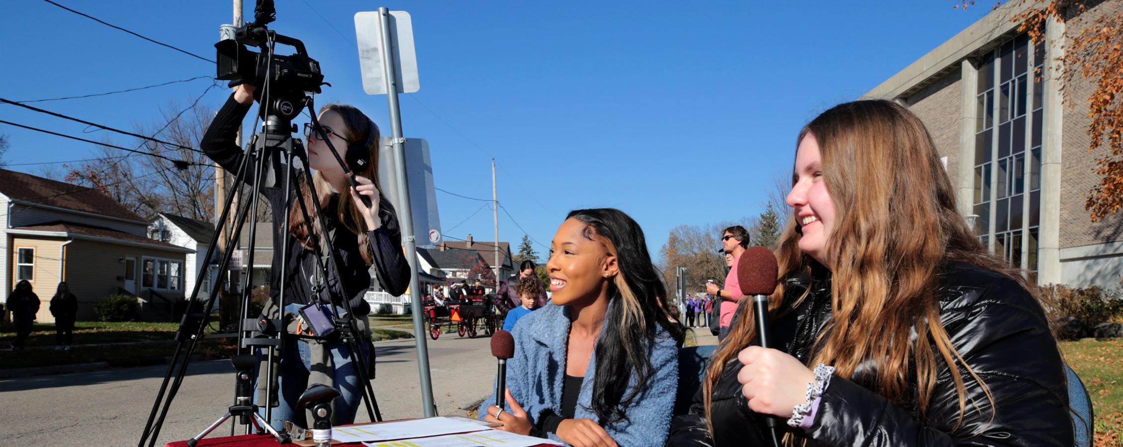 Two students hold microphones and one student works a video camera during a Homecoming parade.