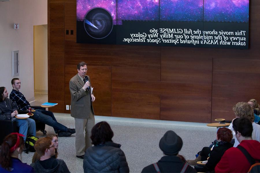 A faculty member speaks to students standing next to a presentation about the Spitzer Space Telescope.