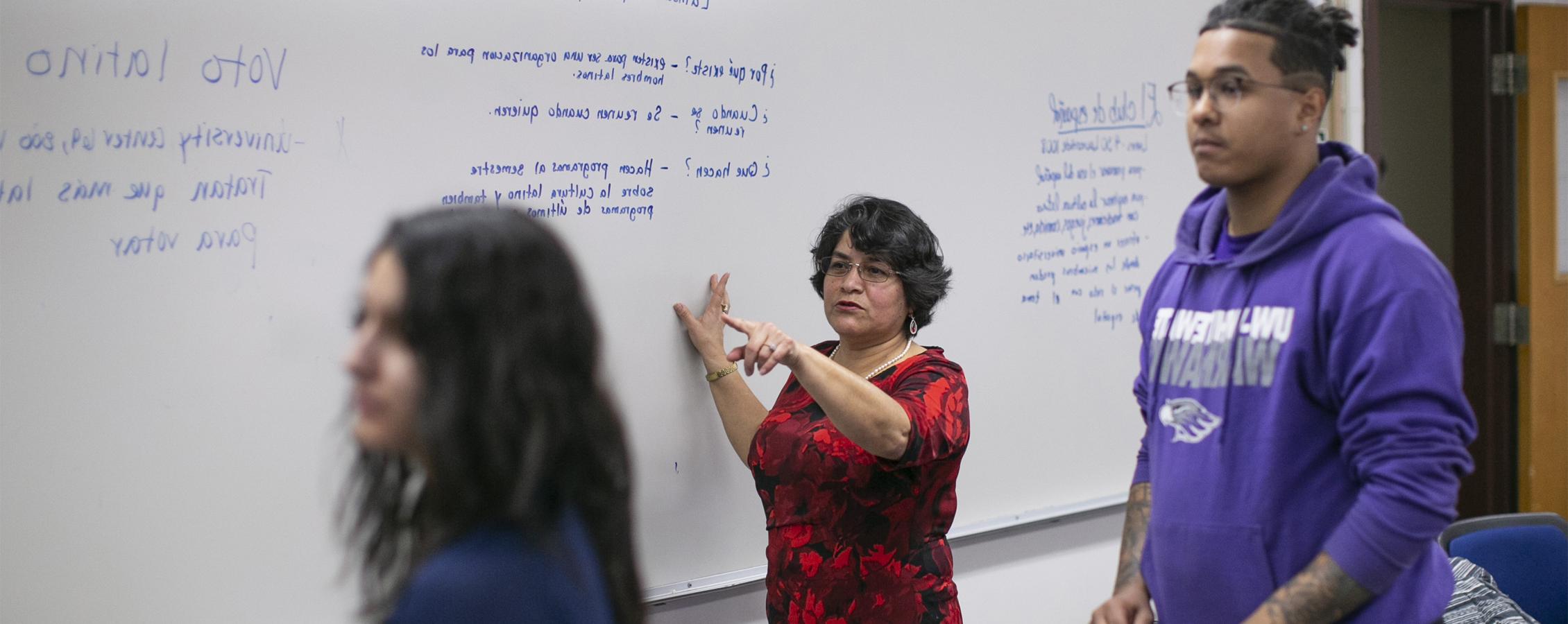 A Spanish faculty member directs students at a whiteboard.