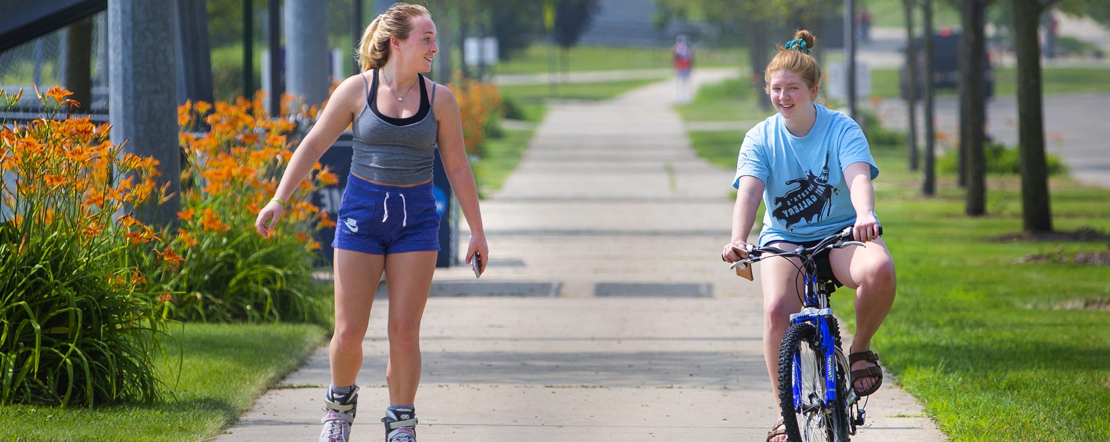 A student rides a bike next to another student rollerblading down a sidewalk.