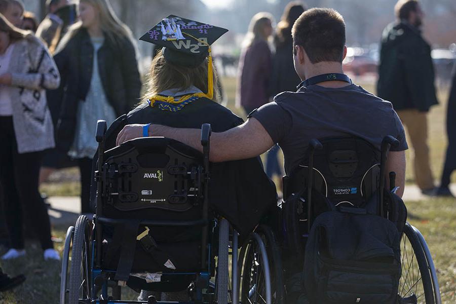 Two people outdoors in wheelchairs with arms around each other at graduation.