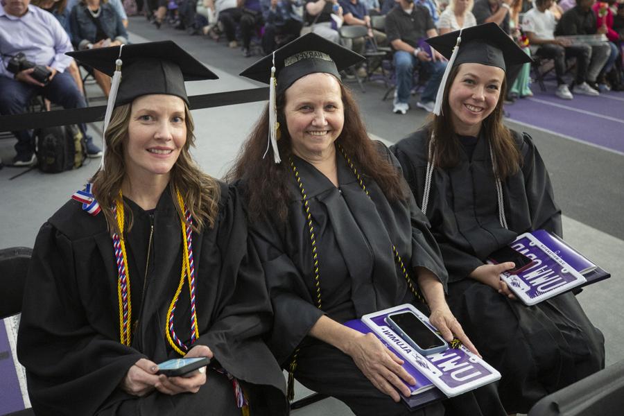 Three adults wearing caps and gowns sit together at graduation.